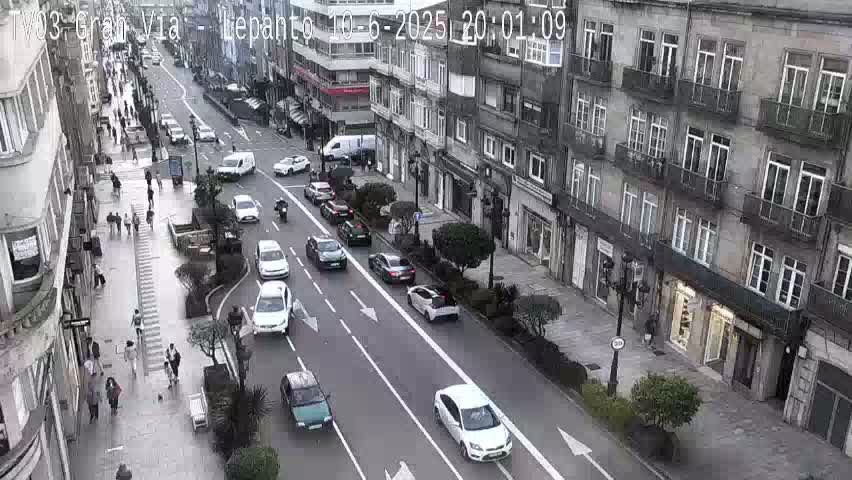 A city street scene on an overcast day shows cars driving in both directions on a multi-lane road, with pedestrians walking on sidewalks lined with multi-story buildings.