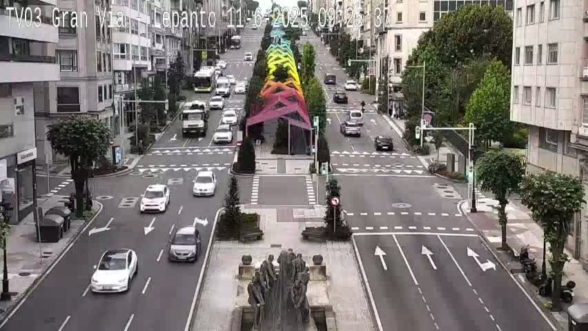 A city street scene on a partly cloudy day shows several cars driving on a wide road that intersects with another road,  with a rainbow-colored structure in the center and a statue at the intersection.