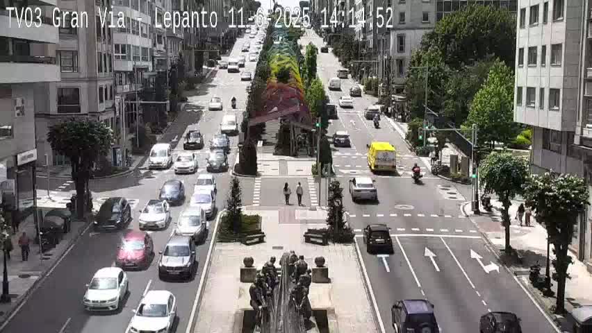 A sunny city street scene shows several cars driving in both directions, with a pedestrian crossing and a statue in the center of the road.