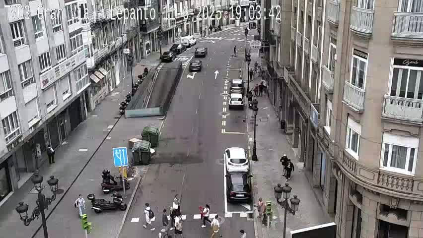A street scene shows several cars and pedestrians on a city street between buildings on a mostly cloudy day.
