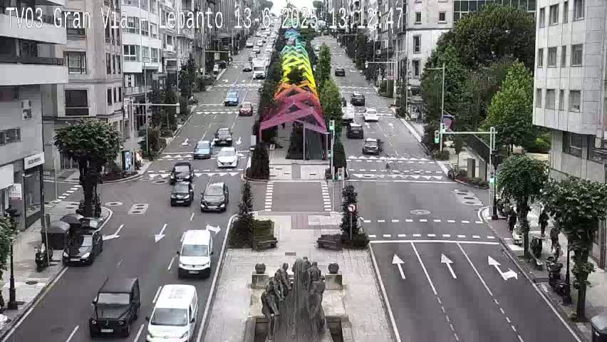 A city street with light traffic, lined with buildings and trees, features a rainbow-colored art installation in the center, under a mostly sunny sky.