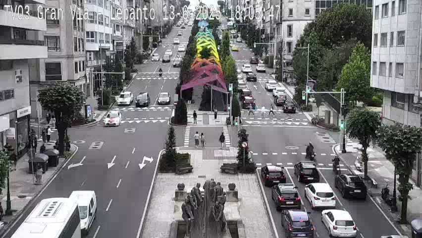 A city street scene on an overcast day shows cars driving on a multi-lane road that intersects with another road, pedestrians walking on sidewalks and in crosswalks, and a large sculpture in the center of the intersection.