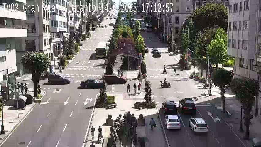 A sunny city street intersection with several cars and pedestrians, and a central median with a sculpture.