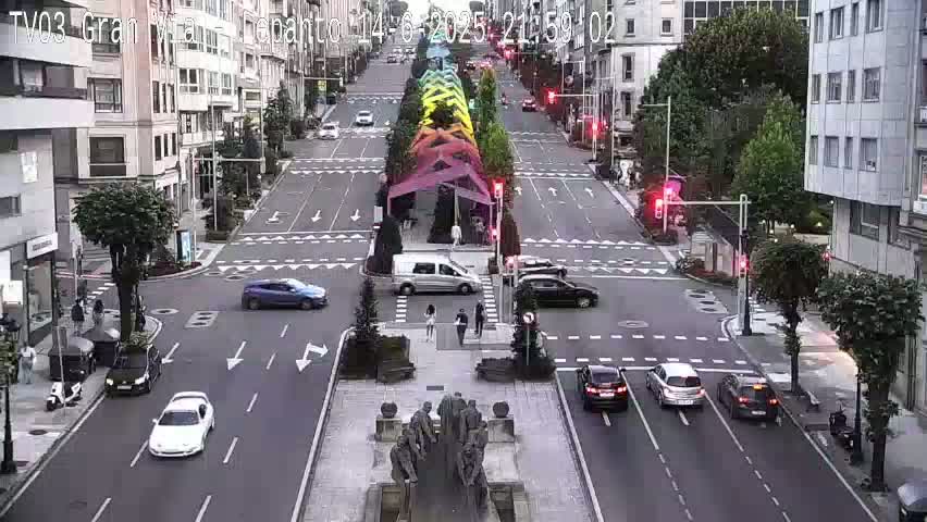 A sunny city street intersection with several cars and pedestrians, and a central median with a sculpture.