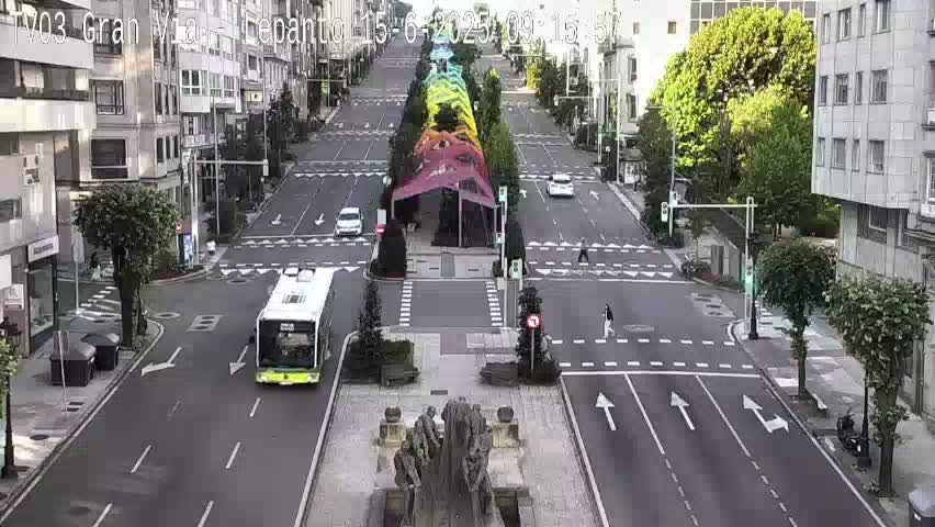 A mostly empty city street with a bus and several cars, a central monument, and buildings lining both sides, under a clear sky.