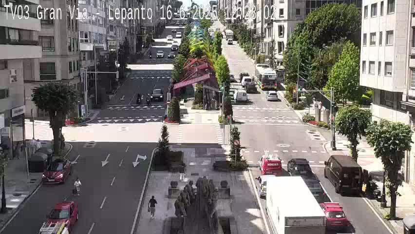 A sunny city street scene shows several cars and pedestrians, with a large sculpture in the center of the road and buildings lining both sides of the street.