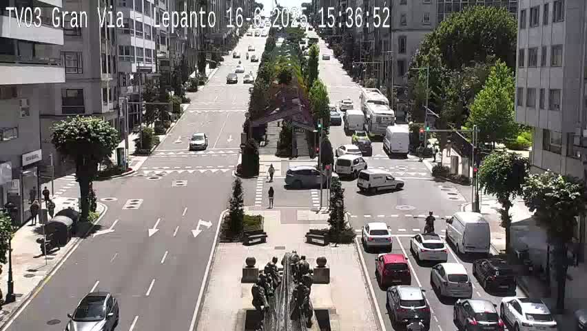 A sunny city street scene shows several cars moving along a wide road with a median featuring a fountain and sculptures, and pedestrians crossing at intersections.
