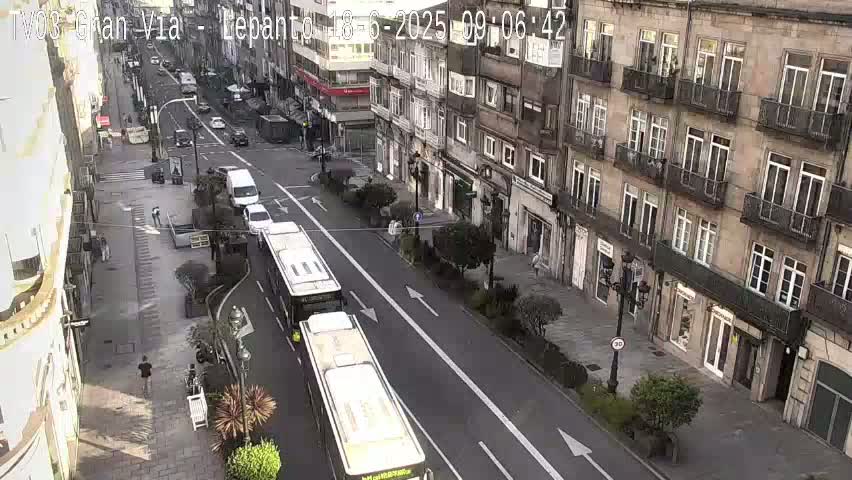 A city street scene on a partly sunny day shows several buses and cars moving along a multi-lane road that passes in front of tall buildings with balconies.