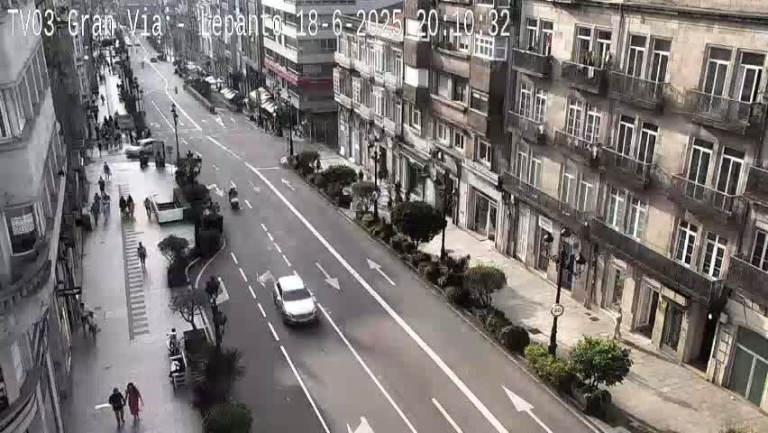 A sunny day on a city street shows vehicles driving and pedestrians walking past multi-story buildings with balconies.