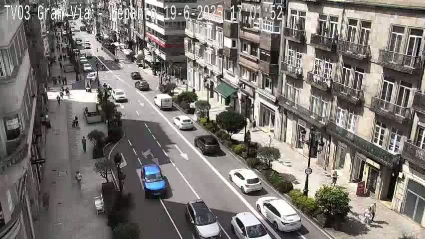 A sunny day on a city street shows several cars driving in both directions past multi-story buildings with balconies and pedestrians walking on sidewalks.