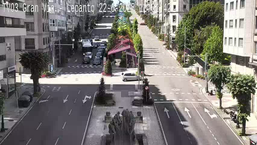 A sunny city street scene shows several cars driving on a road that passes by buildings and a central plaza with a statue.