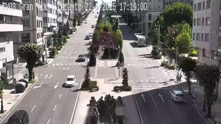 A sunny day on a city street shows several cars driving on a wide road that intersects with another street, with buildings and trees lining the street.