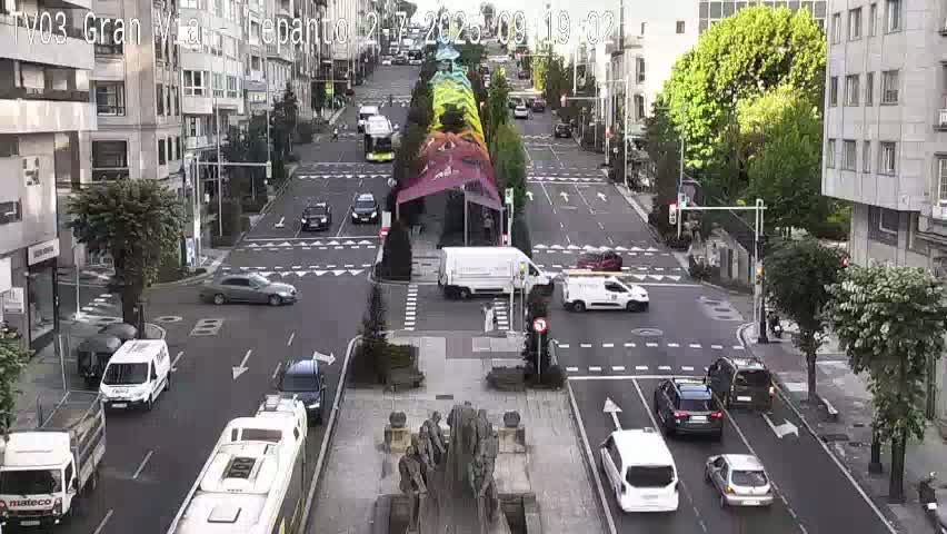 A sunny city street scene shows several cars at an intersection near a statue and a colorful street installation.