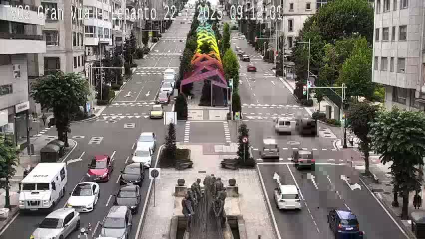 A city street scene with multiple cars driving in both directions, a large statue in the center of the intersection, and a rainbow-colored structure in the middle of the street, under an overcast sky.