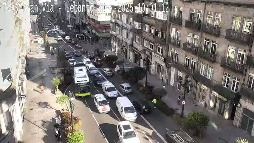 A city street scene on a sunny day shows numerous vehicles moving along a multi-lane road flanked by tall buildings.