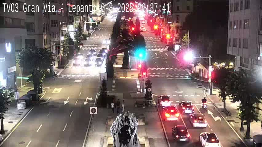 A nighttime, high-angle view shows a multi-lane road intersection with cars driving in both directions, a statue in the center, and buildings lining the street.