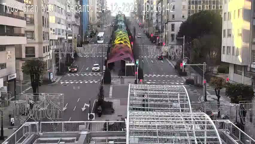 An overhead view captures a bustling city street, lined with multi-story buildings and adorned with decorative overhead light structures, featuring vehicles on the wet road and pedestrians on the sidewalks, all under cloudy skies suggesting recent rain.