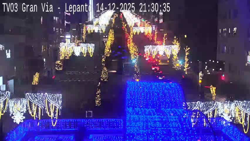 An overhead view captures a bustling city street, lined with multi-story buildings and adorned with decorative overhead light structures, featuring vehicles on the wet road and pedestrians on the sidewalks, all under cloudy skies suggesting recent rain.