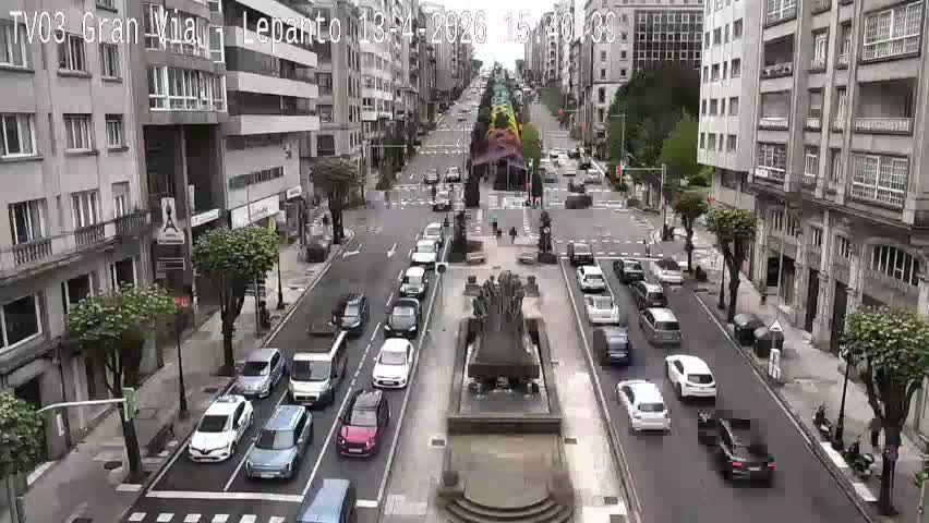 An overhead view captures a bustling city street, lined with multi-story buildings and adorned with decorative overhead light structures, featuring vehicles on the wet road and pedestrians on the sidewalks, all under cloudy skies suggesting recent rain.