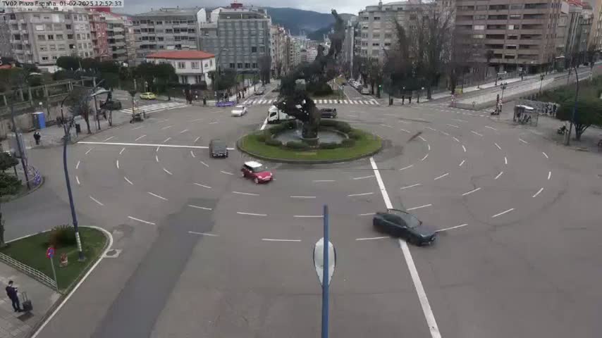 A roundabout with several cars and a large, abstract sculpture in the center, under an overcast sky.