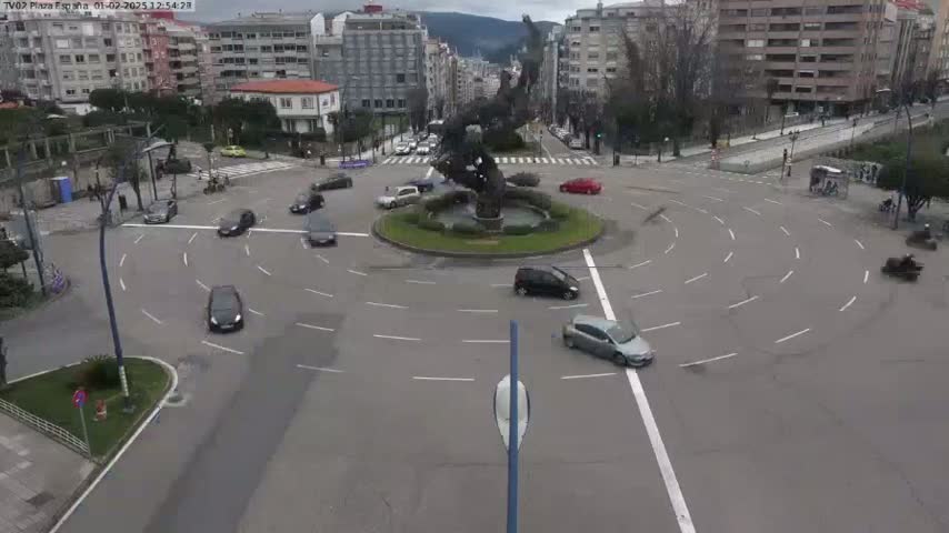 A roundabout with several cars driving around a central fountain and sculpture, under an overcast sky.