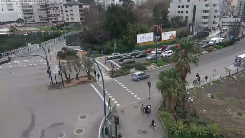 A street intersection with several cars and pedestrians, surrounded by buildings and greenery under a partly cloudy sky.