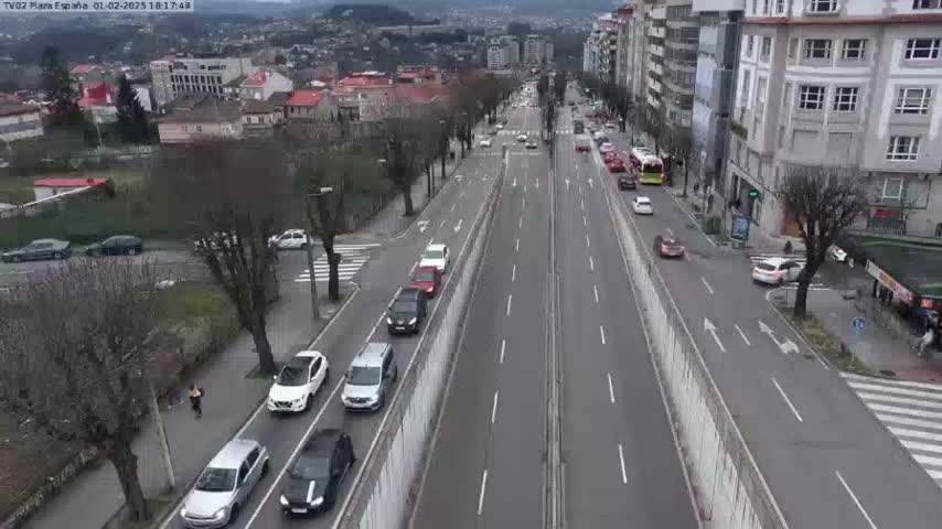 A high-angle, long shot of a multi-lane road with moderate traffic and buildings on either side under an overcast sky.