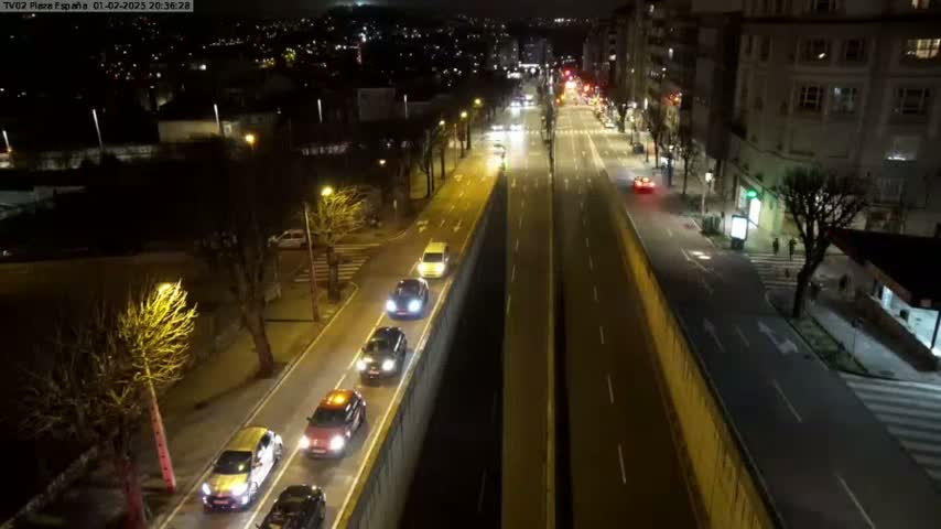 A nighttime aerial view shows several cars driving along a divided highway, flanked by buildings and trees under clear skies.