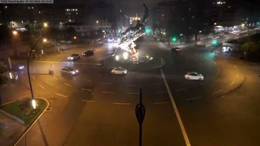 A nighttime, high-angle view shows several cars navigating a roundabout centered around a sculpture, under slightly overcast conditions.