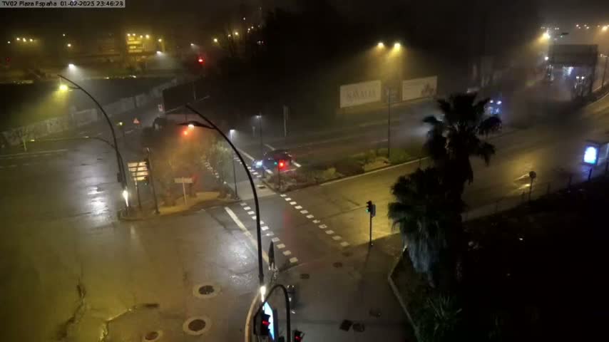 A nighttime, foggy view of a wet road intersection with sparse traffic and streetlights illuminating the scene.