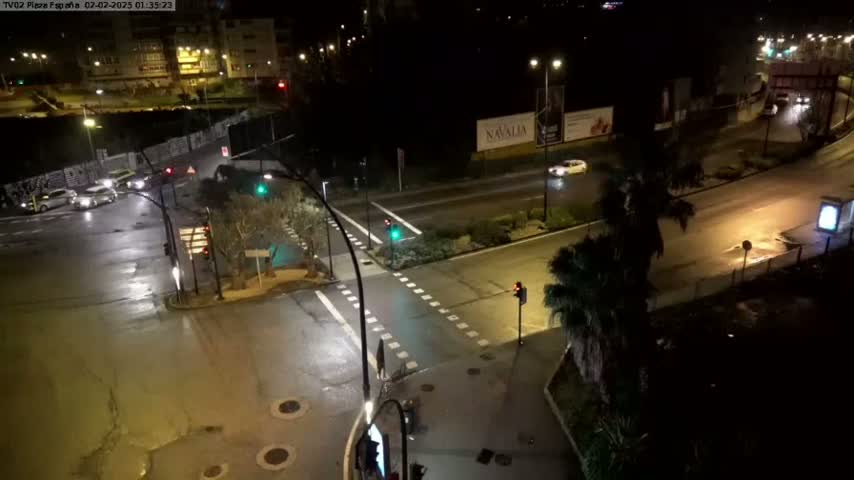 A wet, nighttime street intersection with several cars and traffic lights, surrounded by buildings and some vegetation.