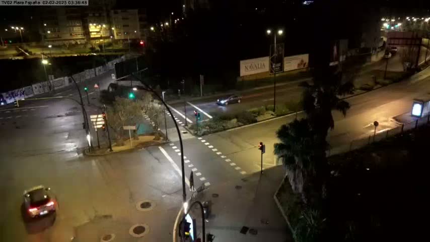 A nighttime, high-angle view of a wet, multi-lane intersection with several vehicles and streetlights, surrounded by buildings and palm trees.
