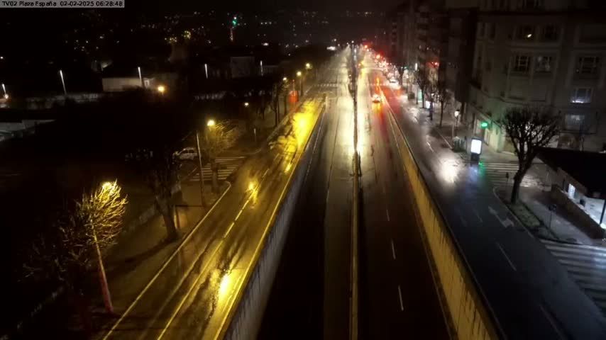 A rain-slicked, multi-lane road is illuminated by streetlights at night, flanked by buildings and trees.
