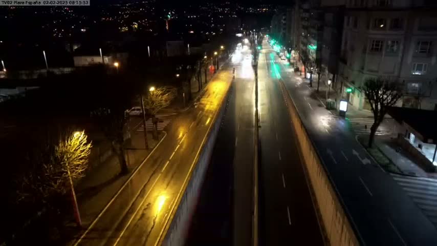 A wet, nighttime view of a mostly empty multi-lane road, bordered by buildings and trees, with streetlights illuminating the scene.
