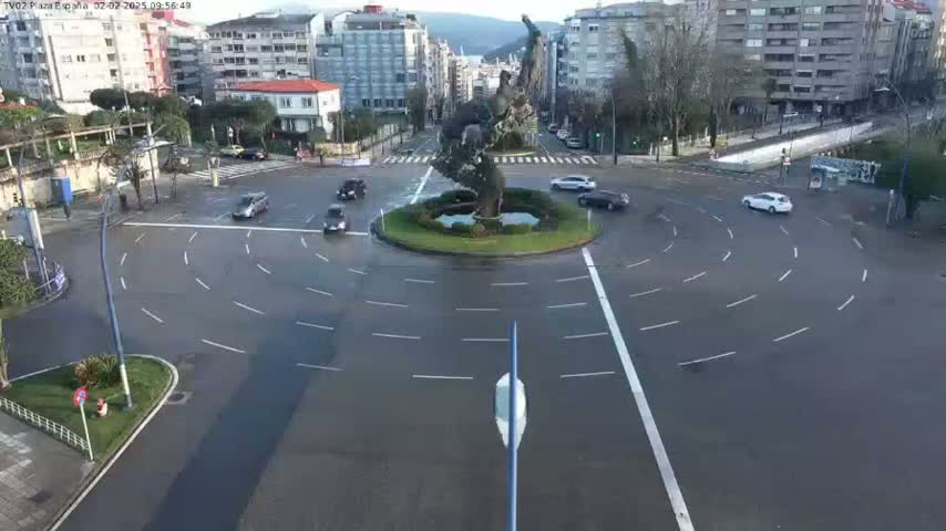 A wet roundabout in a city is surrounded by buildings and a few cars are navigating the circular lanes.