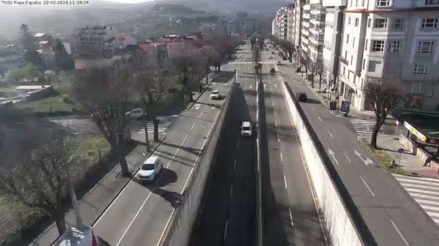 A sunny day reveals a multi-lane road cutting through a city, flanked by buildings and trees, with a few cars visible.