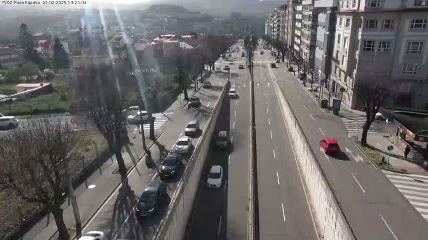 A sunny day shows several cars driving on a multi-lane road that is flanked by buildings and trees.