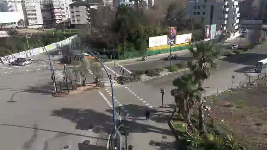 A sunny day shows a street intersection with several cars and pedestrians, surrounded by buildings and palm trees.