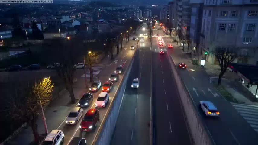 A nighttime, high-angle view of a multi-lane road with moderate traffic, lined with buildings and trees.
