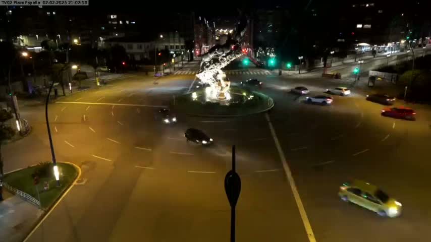 At night, several cars are driving around a roundabout centered on a large sculpture, under clear skies.
