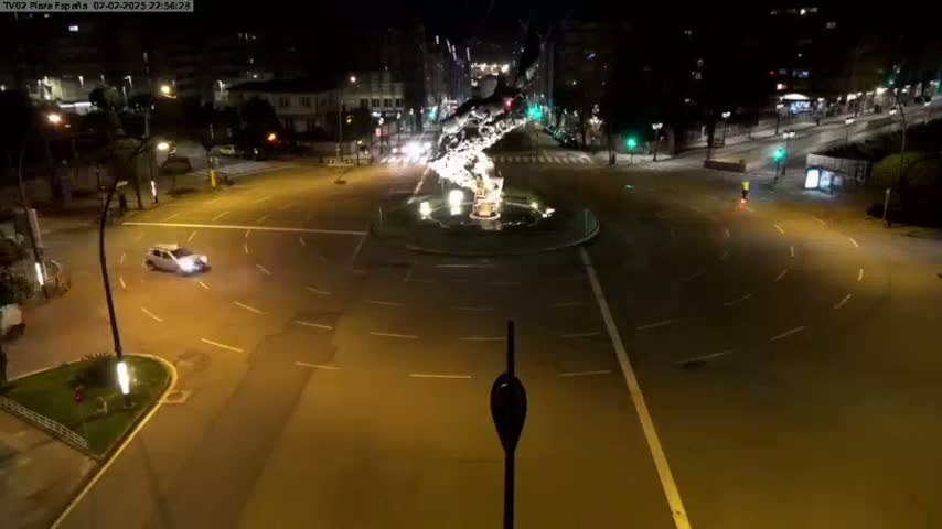 A nighttime aerial view shows a traffic circle with a few cars and illuminated streets.
