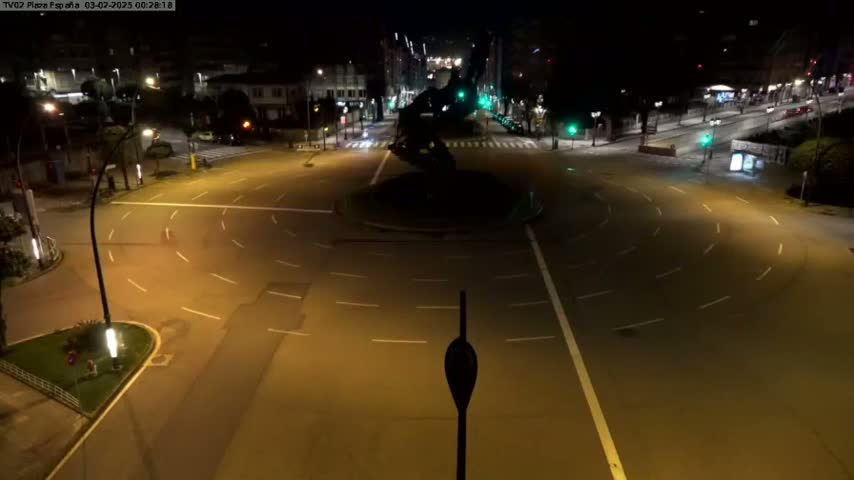 A nighttime, high-angle view of a mostly empty roundabout with a dark statue in the center, surrounded by city streets and buildings.