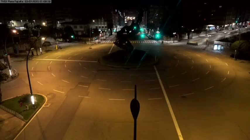 A mostly empty roundabout at night is illuminated by streetlights, with buildings visible in the background.