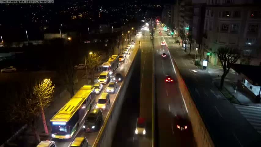 A nighttime, high-angle view of a city street with multiple cars and buses traveling in both directions, alongside a wall separating a road and a sidewalk.