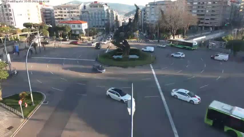 A roundabout with several cars and buses, a statue in the center, and buildings in the background under a partly sunny sky.