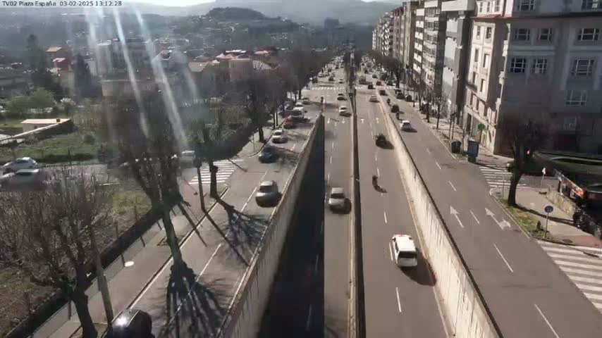 A sunny day overlooking a highway with several cars driving in both directions, flanked by buildings and trees.