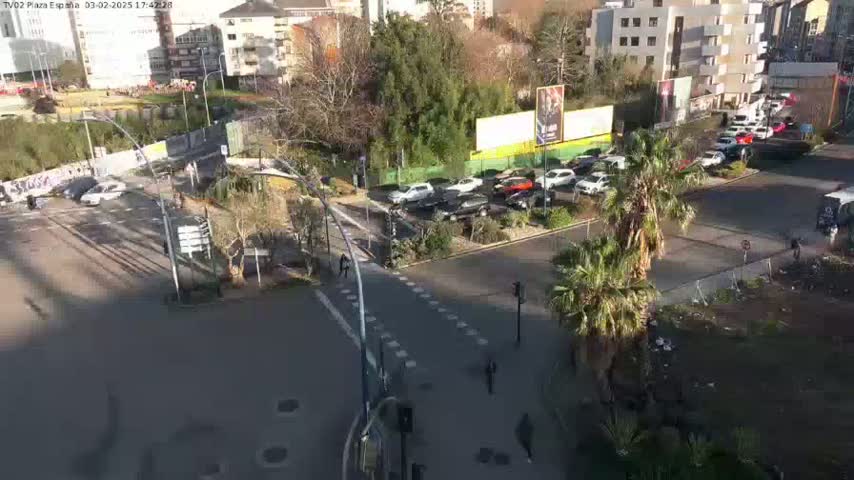 A sunny day shows an aerial view of a city intersection with cars stopped at a crosswalk and pedestrians walking near palm trees and buildings.