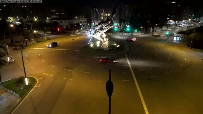 A nighttime aerial view of a roundabout with several cars driving around a central sculpture, under clear skies.