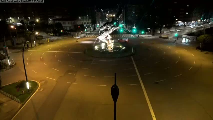 A nighttime, overhead view of a mostly empty roundabout with a sculpture in the center, surrounded by buildings and streetlights.