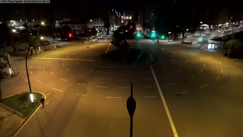 A nighttime, high-angle view shows a mostly empty roundabout with a dark statue in the center, illuminated by streetlights.